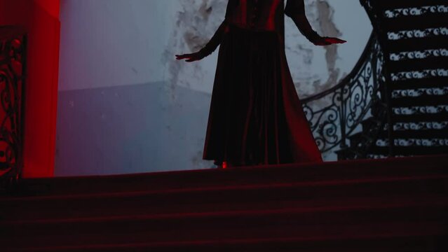 a goth sexy woman in a dark halloween outfit moving confident and pose. The hallway is brightened by red lamps. It's nighttime indoors.