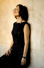 A beautiful young brunette in an evening dress stands relaxed against the wall