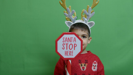 Adorable kid with double headband christmas deer antlers showing sign santa stop here. Isolated on...