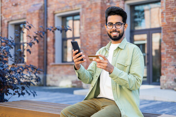 Portrait of a happy young Hindu man using a credit card and phone, sitting outside on a bench and...