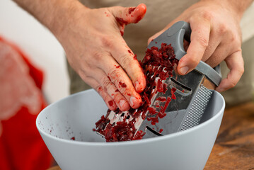 Close-up of hands with red stains using a grater to shred beetroot into a white bowl, healthy cooking concept.