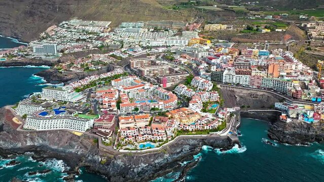 Aerial view of a luxury resort and colourful hotels with swimming pools in Tenerife. Whitewashed houses on a rugged hillside that tumbles down to a tiny black sandy beach in Puerto de Santiago.
