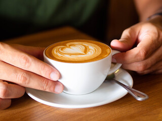 A man hands wrapping around a white cup of hot coffee latte drink on wooden table of coffee shop. Sitting in cafe and carefully holding cappuccino cup with heart shape latte art. Close up milk foam