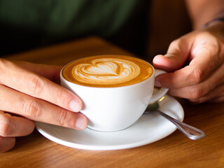A man hands wrapping around a white cup of hot coffee latte drink on wooden table of coffee shop....