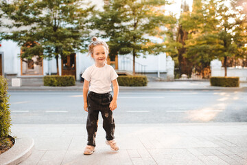 Smiling child girl 4-5 year old wear trendy clothes walking in city street outdoor. Looking at camera. Childhood.