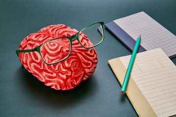 Representation of a brain with glasses and a notebook and pencil on a dark-colored table.