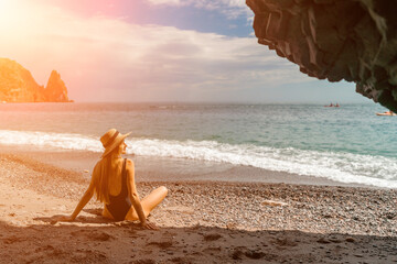 Woman travel sea. View of a woman in a black swimsuit from a sea cave Attractive woman enjoying the sea air sits on the beach and looks at the sea. Behind her are rocks and the sea - Powered by Adobe