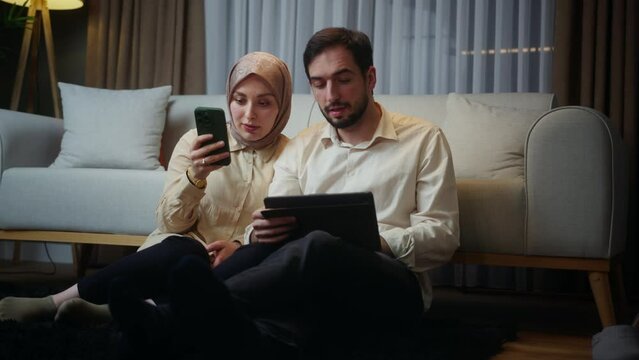 Young married couple leaning against the sofa sitting on floor, woman using smartphone, husband using a tablet device, spending time on internet, using app at home at night