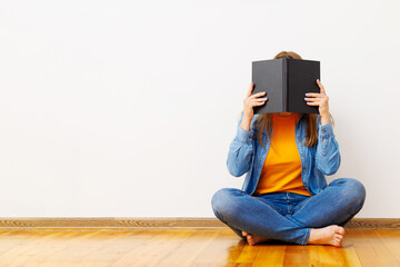 A young woman covers her face with a book as she sits on floor against the wall