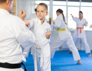 Fototapeta na wymiar Boy in kimono and belt practicing karate punch block during group martial arts lesson in gym, accompanied by trainer