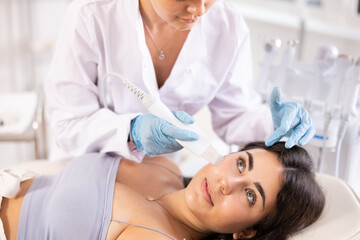 Young brunette undergoing hardware cosmetic treatment for facial care in aesthetic medicine clinic,...