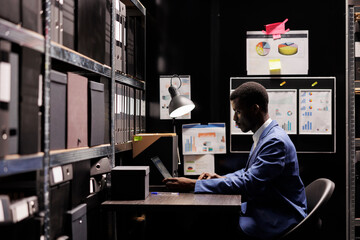African american police officer analyzing investigation files, working overtime at confidential case in arhive room. Private detective checking victim evidence, planning strategy to catch suspect.