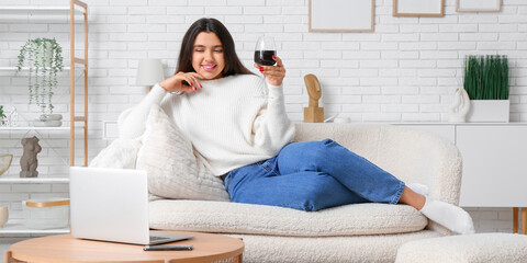 Young woman with laptop and glass of wine sitting on sofa at home