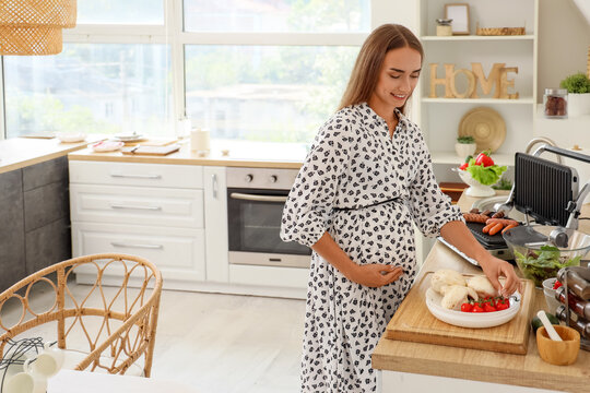 Beautiful young pregnant woman preparing vegetables near electric grill with sausages in kitchen