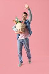 Young man with grocery bag waving hand on pink background