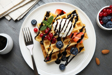Delicious Belgian waffles with ice cream, berries and chocolate sauce served on grey textured table, flat lay