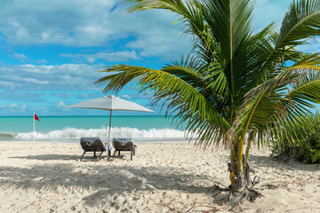 Two lounge chairs covered with towels, an umbrella, a palm tree and a warning red flag on a beach