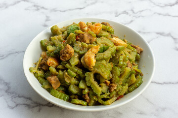 Stir-fried bitter gourd or bitter melon mixed with shrimp and tempeh on white plate, an easy-to-cook dish with healthy benefits A typical main course of Asian food eaten with rice. Top view