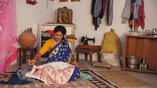 A middle-aged woman in her late forties is folding the clothes kept in a heap - household chores. A poor Indian lady from an Indian village - rural household  BPL  below the poverty line family  si...