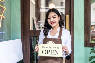 Business owner happy beautiful young asian woman in apron looking at camera, we're open sign on front door smiling welcoming clients to new cafe, People and start-up concept.