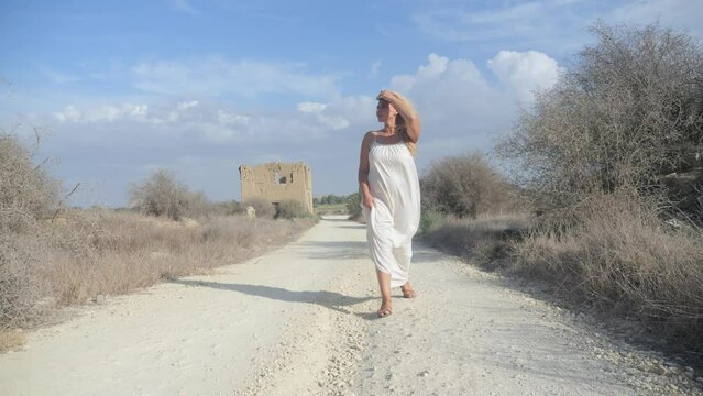 Wide shot of a woman in a white dress searches for something down a deserted dirt path with ruins in the background.
