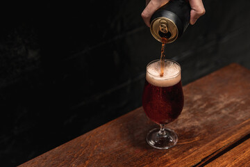 Hand pouring cold black beer from mockup can to wine glass. Summer refreshing drink. - obrazy, fototapety, plakaty