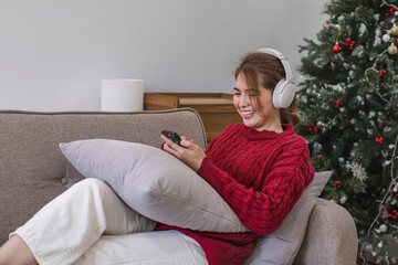 Asian woman sitting on sofa in living room using smartphone and listening to music on headphones.