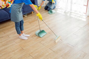 Crop image of a young professional cleaning service women worker working in the house. Girls housekeeper sweeps broomsticks on the wooden floor and cleaning under the sofa. Cleaner and chore concept - obrazy, fototapety, plakaty