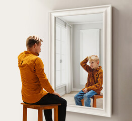 Man in his 30s sitting and looking in mirror with reflection his reflection as a child. Childhood...