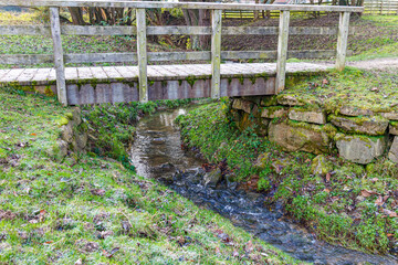Small wooden pedestrian bridge over a narrow stream between green grass, water flowing over stones,...