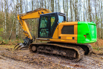 Side angle perspective of grapple excavator on muddy ground, bare trees against blue sky in...
