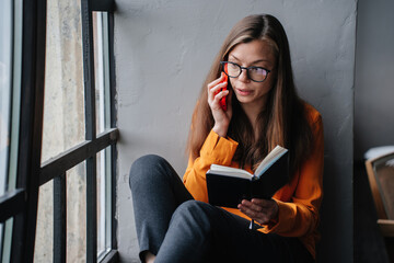 Intellectual young woman in a vivid orange blouse deeply engrossed in a book while sitting by a...