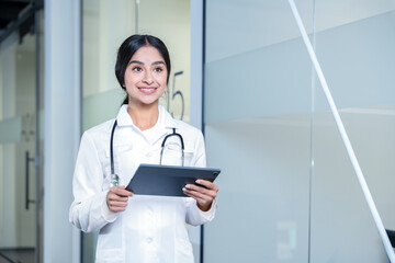 Young female doctor in white coat and stethoscope confidently holds a tablet, portraying professionalism and dedication to patient care in a bright, clean medical setting.