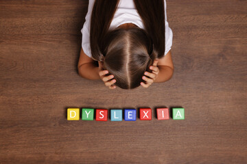 Little girl covering head with hands and cubes with word Dyslexia at wooden table, top view - obrazy, fototapety, plakaty
