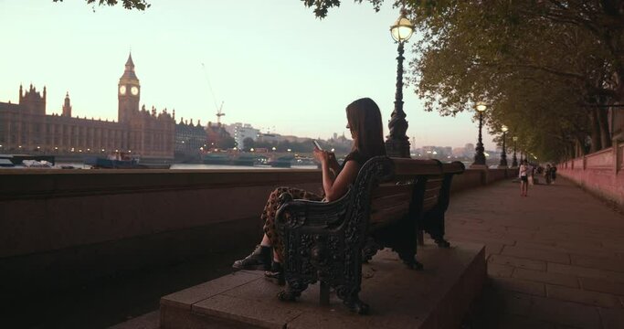 Happy local beautiful female chats via phone, sitting on bench with the view of London famous Big Ben in the evening.