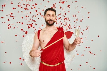 bearded man in cupid costume with heart-shaped arrow and champagne under red confetti on grey
