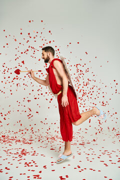 man in cupid costume posing with heart-shaped arrow under red confetti on grey, Saint Valentines day