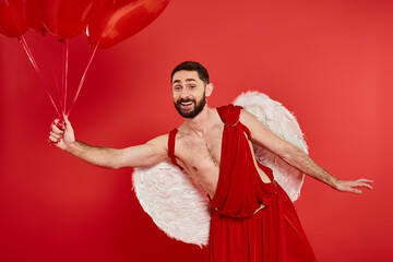 smiling bearded man in cupid costume posing with heart-shaped balloons on red, st valentines day