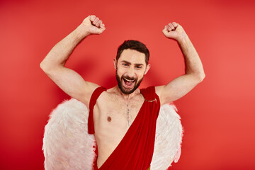 excited bearded cupid man rejoicing and showing win gesture with raised hands on red backdrop