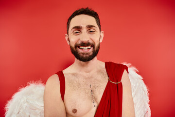 portrait of cheerful bearded man dressed as cupid smiling at camera on red, st valentines party