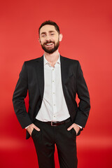 joyful businessman in black elegant suit standing with hands in pockets and smiling at camera on red