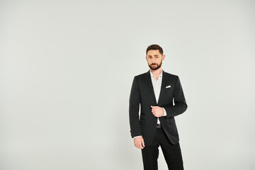 Obraz na płótnie Canvas good-looking bearded businessman in black stylish suit standing and looking away on grey backdrop