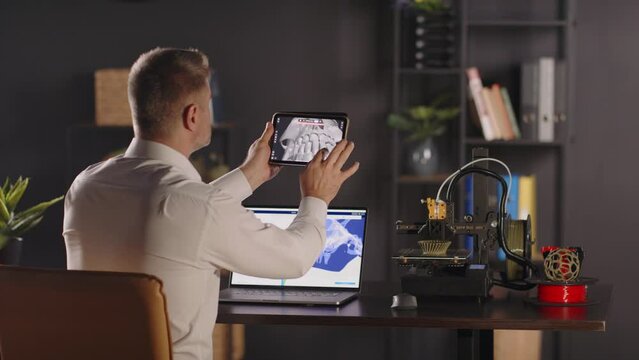 A man graphic designer sits back by the table, uses a tablet to view 3d model of a human skull and teeth. A work laptop for creating digital sketches. The concept of bioprinting organs for transplants