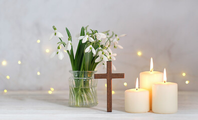 Wooden Christian cross, snowdrops flowers and candles on table, light abstract background. Religious church holiday. symbol of faith in God, Christianity Feast. Christmas, Easter, Palm Sunday holidays