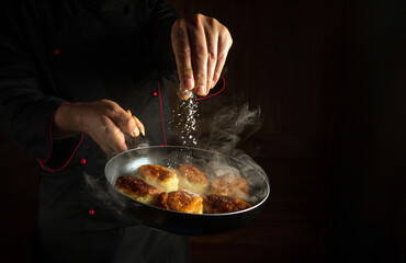 A cook salts hot pancakes in a frying pan while frying. The idea of ​​cooking pancakes in the kitchen. Dark space for advertising