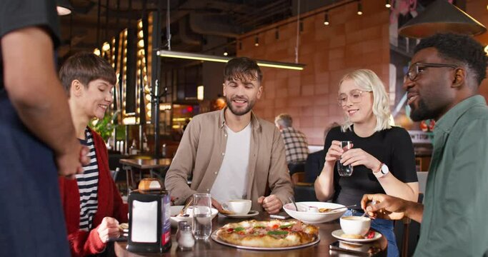 Zooming out from positive company sitting at table full of tasty dish. Caring waiter in apron speaking with his clients in cafe. Serving people in restaurant. Concept of customers.