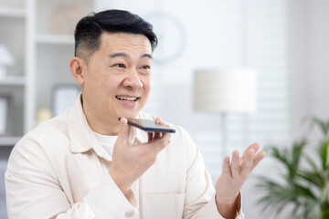 Close-up photo of a young smiling Asian man sitting at home talking on the phone through the speakerphone, recording the conversation, asking in the application.