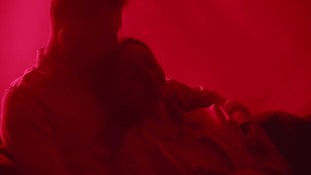 Sweet couple sitting in bright neon light. Lovely family lying at couch together indoors. Two unrecognizable people embrace kisses on colourful light background. Passionate man kissing girlfriend