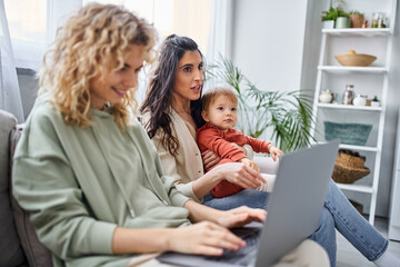 focus on brunette woman with baby girl sitting on sofa near her blurred partner with laptop, family