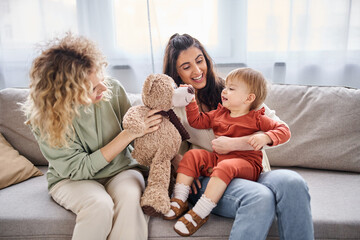 attractive jolly lesbian couple with their toddler daughter and teddy bear on sofa, family concept